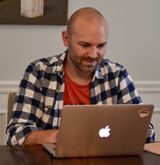 Chris is wearing a red shirt and a black and white plaid long shirt, sitting at a table and typing on a Macbook. The Apple logo is illuminated on the back of the laptop.