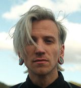 Close-up photo of Henry’s face, with chin-length light colored hair and a neutral facial expression. A cloudy blue sky sits in the background.